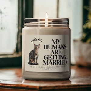May include: A white candle with a black and white label that says "smells like my humans are getting married". The label also features a picture of a tabby cat. The candle is on a wooden surface.
