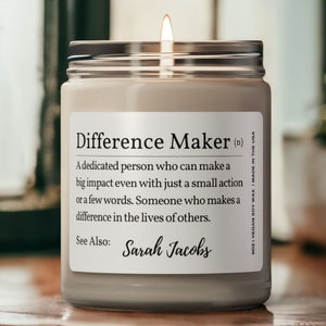 K&ouml;nnte beinhalten: Eine beige Kerze in einem Glasgef&auml;&szlig; mit einem silbernen Deckel. Die Kerze hat ein Etikett mit der Aufschrift "Difference Maker (n)" und definiert den Begriff als "Eine engagierte Person, die auch mit einer kleinen Handlung oder wenigen Worten einen gro&szlig;en Einfluss haben kann. Jemand, der einen Unterschied im Leben anderer macht." Das Etikett enth&auml;lt auch den Text "Siehe auch: Sarah Jacobs".