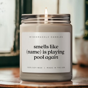 May include: A white candle with a black label that reads "WISECRACKLE CANDLES smells like (name) is playing pool again 100% SOY WAX | MADE IN THE USA".
