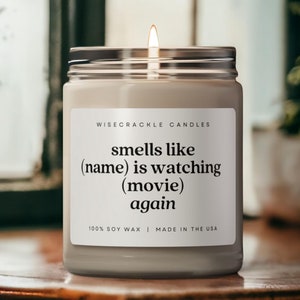 May include: A white candle with a black label that reads "WISECRACKLE CANDLES smells like (name) is watching (movie) again 100% SOY WAX | MADE IN THE USA".