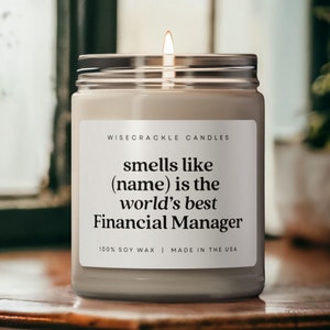 May include: A white candle with a black label that reads "WISECRACKLE CANDLES smells like (name) is the world's best Financial Manager 100% SOY WAX | MADE IN THE USA".