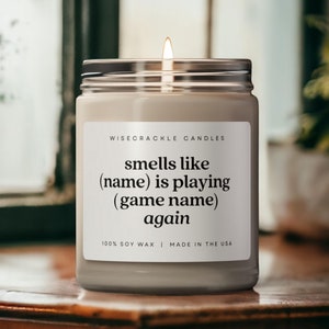 Könnte beinhalten: Eine weiße Kerze mit einem schwarzen Etikett, auf dem steht: "WISECRACKLE CANDLES smells like (name) is playing (game name) again 100% SOY WAX | MADE IN THE USA".
