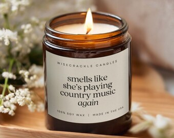 May include: A lit soy wax candle in a brown glass jar. The label reads "smells like she's playing country music again." The candle is on a wooden tray, surrounded by white flowers. The text "WISECRACKLE CANDLES" and "100% SOY WAX | MADE IN THE USA" are also visible.
