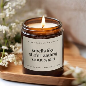Pode incluir: Uma vela acesa num frasco de vidro castanho com uma etiqueta branca. A etiqueta diz "smells like she's reading smut again". A vela está numa bandeja de madeira, com flores brancas ao fundo. O texto "100% SOY WAX I MADE IN THE USA" também é visível.