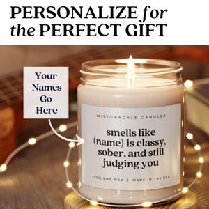May include: A white candle with a black label that reads "smells like (name) is classy, sober, and still judging you". The label also says "Wisecrackle Candles" and "100% Soy Wax | Made in the USA".
