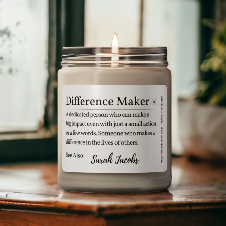 K&ouml;nnte beinhalten: Eine beige Kerze in einem Glasgef&auml;&szlig; mit einem silbernen Deckel. Die Kerze hat ein Etikett mit der Aufschrift "Difference Maker (n)" und definiert den Begriff als "Eine engagierte Person, die auch mit einer kleinen Handlung oder wenigen Worten einen gro&szlig;en Einfluss haben kann. Jemand, der einen Unterschied im Leben anderer macht." Das Etikett enth&auml;lt auch den Text "Siehe auch: Sarah Jacobs".