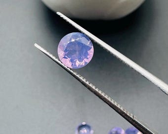 Könnte beinhalten: Eine Sammlung kleiner, runder, lavendelfarbener Edelsteine. Ein Edelstein wird von einer silbernen Pinzette gehalten, während andere auf einer dunklen Oberfläche verstreut sind. Ein weißer Keramiktopf mit einer grünen Pflanze befindet sich im Hintergrund.