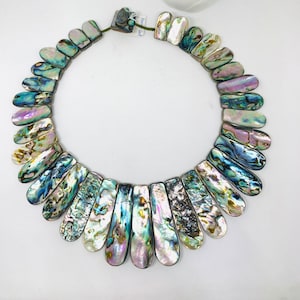 Peut inclure: Un collier composé de pièces de coquille d'ormeau irisées dans des tons de bleu, vert et violet. Les pièces sont en forme de larmes et sont disposées en forme d'éventail.