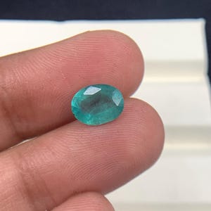 May include: An oval-cut, teal gemstone. The gem is held in a hand against a dark background. The stone has facets that catch the light, creating a sparkling effect. This gemstone could be used in jewelry.