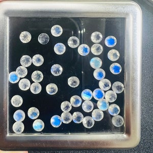 May include: A close-up of a silver box containing a collection of round, iridescent white gemstones. The gemstones are arranged in a random pattern and have a subtle blue sheen.