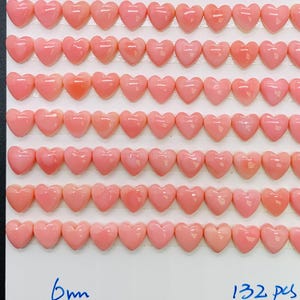 May include: A collection of 132 heart-shaped beads in varying shades of pink, arranged in rows on a white surface. The beads are approximately 6mm in size. The overall arrangement is neat and symmetrical, with a dark background.