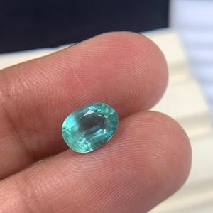 May include: An oval-cut, teal gemstone. The gem is held between fingers, showcasing its polished facets and translucent quality. The stone's color ranges from light to medium teal, reflecting light.