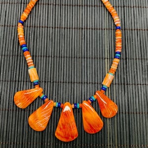 May include: A necklace made of orange, white and blue beads with a pendant of five orange, carved, teardrop-shaped stones.