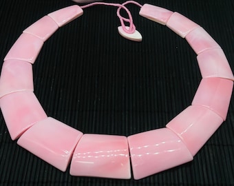 May include: A pink shell necklace with a pink cord closure. The necklace is made of 12 individual shell pieces, each with a slightly different shape and size. The necklace is lying on a black bamboo mat.