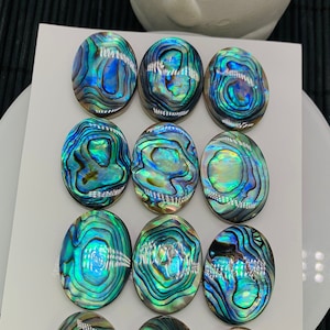 Può includere: Dodici cabochon ovali in madreperla di abalone iridescente, ognuno con un motivo unico di vortici blu, verdi e dorati. I cabochon sono disposti in tre file di quattro su uno sfondo bianco. Il testo "8x25mm" e "12pcs" è scritto nella parte inferiore dell'immagine.
