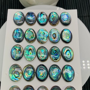 May include: Twenty-five iridescent abalone shell cabochons, each measuring 12x16mm, arranged in rows on a white card. The cabochons have a blue, green, and gold color scheme.