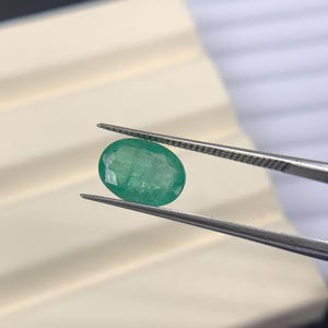 May include: An oval-cut emerald gemstone held by tweezers. The emerald is a vibrant green color and appears to be faceted. The tweezers are silver-colored, and the background is a neutral beige and white.