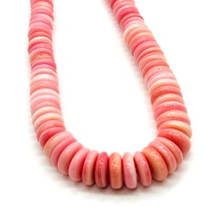 Puede incluir: Un collar de concha rosa con un diseño graduado. El collar presenta cuentas planas y redondas en varios tonos de rosa, desde claro hasta coral oscuro. Las cuentas están ensartadas, creando una apariencia suave y fluida.