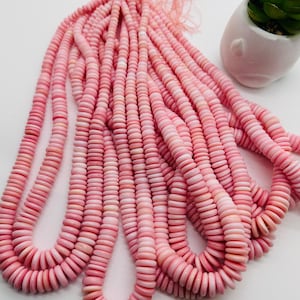 May include: Strands of pink, flat, round beads, likely for jewelry making, are displayed against a white background. The beads vary in shades of pink, from light to darker hues. A small succulent plant in a white pot is in the background.