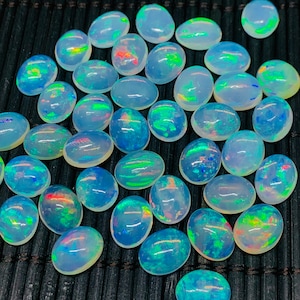 May include: A collection of oval-shaped opals with iridescent color flashes of green, blue, and red. The opals are displayed on a dark, textured surface, showcasing their natural beauty and unique patterns. These gemstones are ideal for jewelry making.