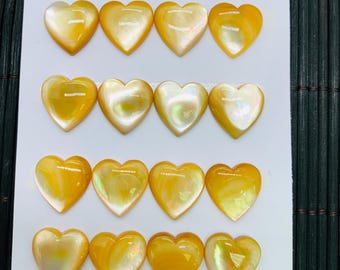 Könnte beinhalten: Zwanzig herzförmige Cabochons in Gelb- und Orangetönen mit schillerndem Glanz. Jedes Herz misst etwa 15 mm. Die Cabochons sind auf einer weißen Karte vor einem dunkelgrünen Hintergrund angeordnet.