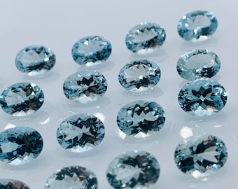 Puede incluir: Una colección de gemas ovaladas de color azul claro. Las gemas están dispuestas sobre una superficie blanca, con algunas hojas verdes en el fondo. Las piedras tienen una superficie pulida y reflectante.