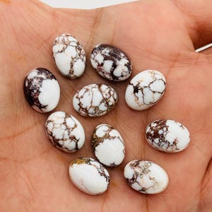 May include: A collection of ten oval-shaped white gemstones with brown and black veining. The stones are smooth and polished, with a marbled appearance. The gemstones are held in a hand, showcasing their size and detail. These stones are ideal for jewelry making.