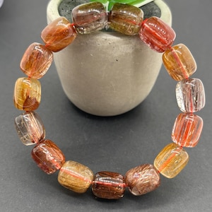May include: A bracelet featuring rectangular, translucent beads with reddish-brown and golden inclusions. The beads are strung together with a red cord, creating a circular shape. The bracelet is displayed against a dark background.