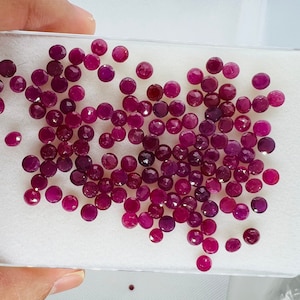 May include: A collection of approximately 70 round, faceted ruby gemstones in various shades of pink and red. The gemstones are loose and scattered on a white surface.