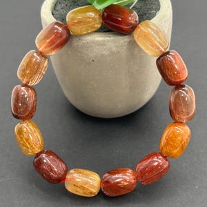 Pode incluir: Uma pulseira de contas ovais em tons de âmbar, laranja e vermelho escuro. As contas são enfiadas, criando um design circular. A pulseira é exibida em uma superfície escura.
