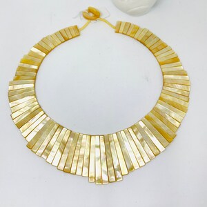 May include: A yellow, iridescent, shell necklace with a fan-shaped design. The necklace is made of many small, rectangular pieces of shell that are arranged in a graduated pattern.