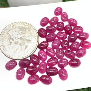 Puede incluir: Una colecci&oacute;n de aproximadamente 30 piedras preciosas de rub&iacute; rosa de forma ovalada. Las piedras preciosas est&aacute;n esparcidas sobre una superficie blanca, con una moneda de veinticinco centavos de d&oacute;lar estadounidense para la escala.
