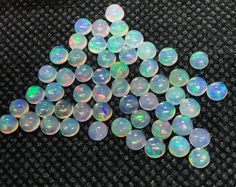 May include: A collection of round, iridescent opal cabochons in various shades of white, with a rainbow of colors reflecting from the surface. The opals are arranged in a triangular shape on a black textured background.