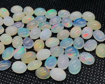 May include: A collection of oval-shaped white opal gemstones with iridescent rainbow colors. The opals are arranged in a loose pile on a black textured surface.