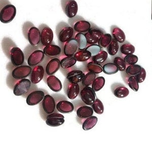 May include: A collection of oval-shaped, translucent, deep red gemstones. The gemstones are scattered on a white surface.