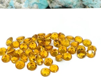 May include: A pile of round, faceted, yellow gemstones. The gemstones are scattered on a white surface.