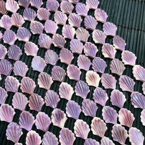May include: A collection of small, seashell-shaped decorative pieces in shades of lavender, purple, and white. The shells are arranged in a grid pattern, with each shell featuring detailed ridges. The background is a dark green, textured surface.