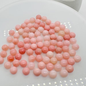 May include: A pile of pink opal cabochons, round and smooth, arranged on a white surface. The cabochons are a light pink color with a slight shimmer.