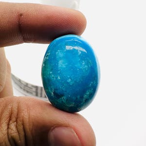 May include: A polished, oval-shaped gemstone in shades of turquoise and teal, held between fingers. The stone has a glossy surface with visible internal patterns. A small succulent in a white pot is in the background.