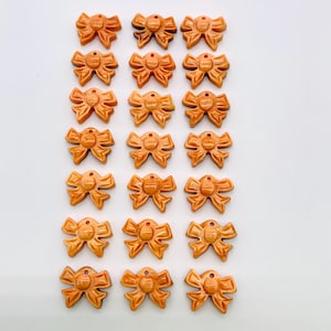 May include: A collection of small, orange-brown bow-shaped charms. Each charm has a detailed bow design with a small hole at the top. The charms are arranged in neat rows against a white background. These charms are suitable for jewelry making or craft projects.