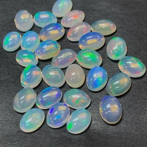 May include: A collection of 20 oval-shaped, white opal gemstones with iridescent rainbow colors. The opals are arranged in a random pattern on a dark gray surface.