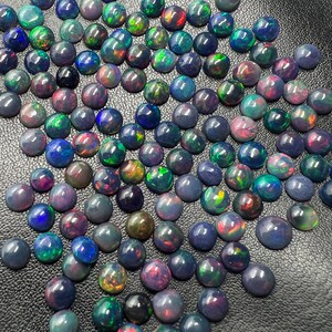 Può includere: Una collezione di cabochon rotondi di opale nero con colori iridescenti, tra cui verde, blu, rosso e viola. Gli opali sono disposti in un motivo sciolto e sovrapposto su una superficie nera.