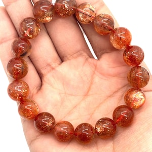 May include: A beaded bracelet featuring large, round, orange-red sunstone beads. The beads have a sparkling, glittery appearance with internal inclusions. The bracelet is strung on a red elastic cord and is displayed on a person's hand.