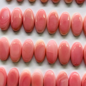 May include: Rows of oval-shaped, polished gemstones in varying shades of pink. The gemstones are arranged neatly on a white surface, showcasing their smooth, glossy texture and subtle color variations. The overall aesthetic is clean and elegant.