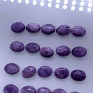 May include: Twenty oval-shaped purple gemstones with a swirling pattern. The stones are arranged in four rows on a white surface.