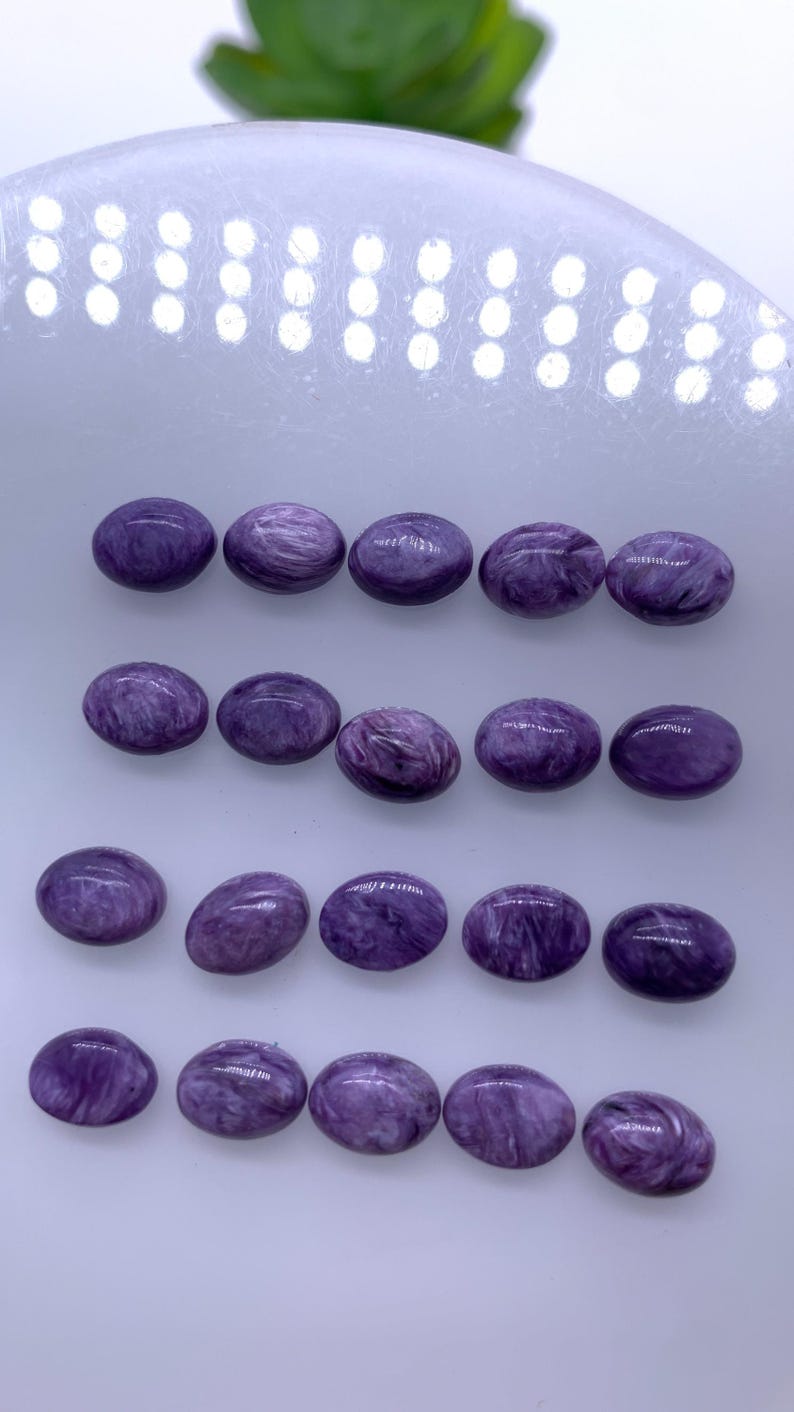 May include: Twenty oval-shaped purple gemstones with a swirling pattern. The stones are arranged in four rows on a white surface.