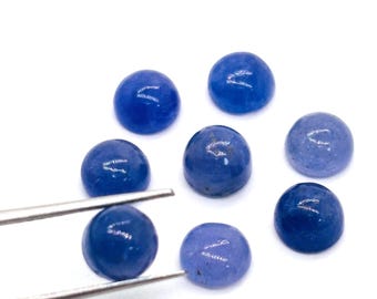 May include: Eight round, polished blue gemstones are displayed against a white background. One gemstone is held by silver tweezers. The gemstones vary in shade, from deep indigo to lighter blues. Ideal for jewellery making.