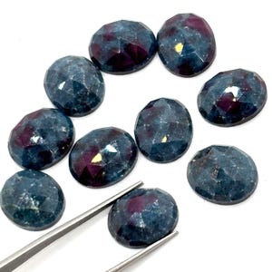 Könnte beinhalten: Eine Sammlung ovaler Edelsteine mit Facettenschliff. Die Steine haben eine dunkelblaue Farbe mit tiefroten und violetten Reflexen. Eine silberne Pinzette hält einen der Steine und zeigt die polierte Oberfläche des Edelsteins.