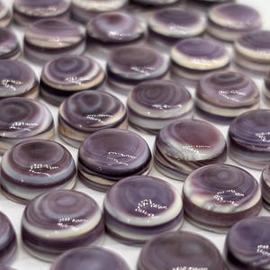 Op de afbeelding: Een close-up van talrijke kleine, ronde cabochons. Ze hebben een wervelend patroon van paars en wit, met een glanzende afwerking. De cabochons zijn dicht bij elkaar gerangschikt op een wit oppervlak.