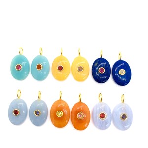 Può includere: Una collezione di pendenti ovali in pietre preziose di vari colori, tra cui blu, giallo e arancione. Ogni pendente presenta una piccola pietra preziosa a contrasto al centro e un'ansa dorata per appenderlo. Sfondo bianco.